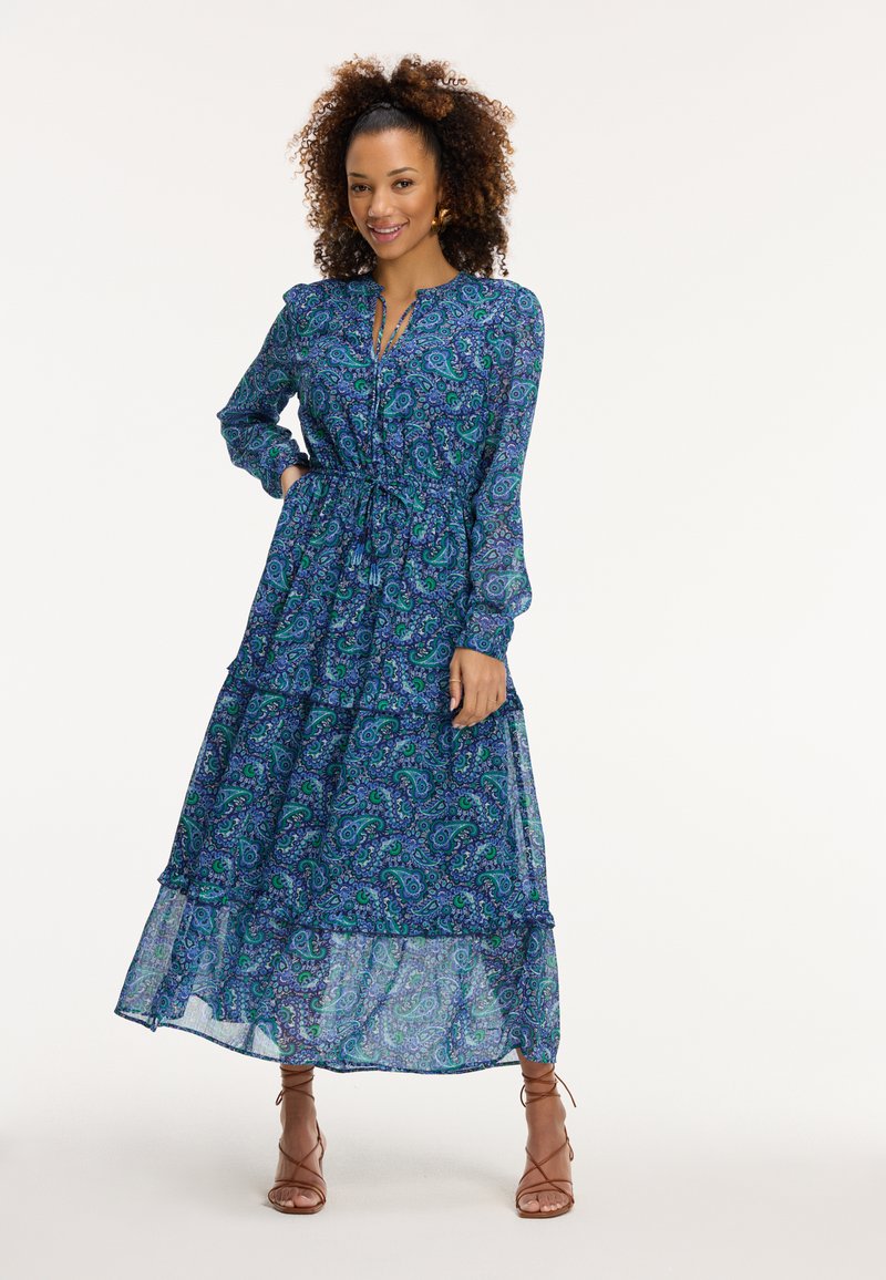 Pitkä sininen paisley-mekko, jossa on kerroksellinen muotoilu, pitkät hihat ja nauhavyötärö. Valmistettu kevyestä, kuviollisesta kankaasta.