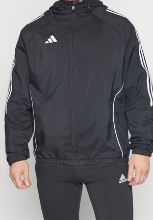 Homme portant une veste de survêtement Adidas noire avec des bandes blanches sur les manches et un pantalon Adidas noir assorti avec un logo sur la cuisse.