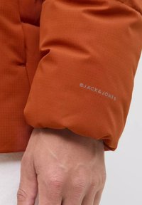 Orange puffer jacket sleeve with a textured finish and visible stitching. Includes a gray logo "JACK & JONES" near the cuff.