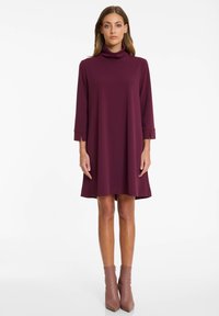 Robe bordeaux avec un col haut, des manches trois-quarts et une silhouette évasée, présentant des lignes épurées et une texture de tissu lisse.