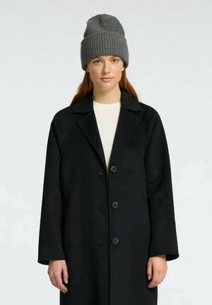 Manteau en laine noire avec un col cranté, comportant quatre boutons à l'avant. Porté sur un haut blanc, associé à un bonnet en maille gris.