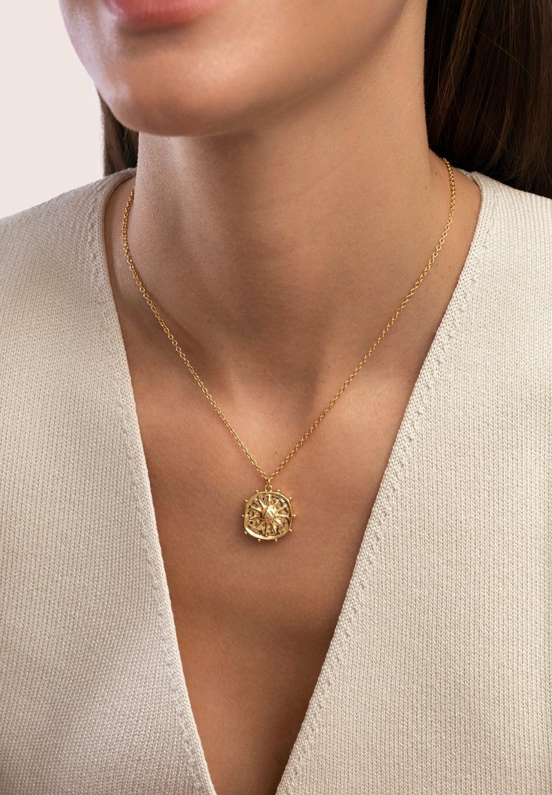 Collier en pendentif doré avec un design complexe, présentant une surface texturée, suspendu à une chaîne délicate, porté sur un haut beige.