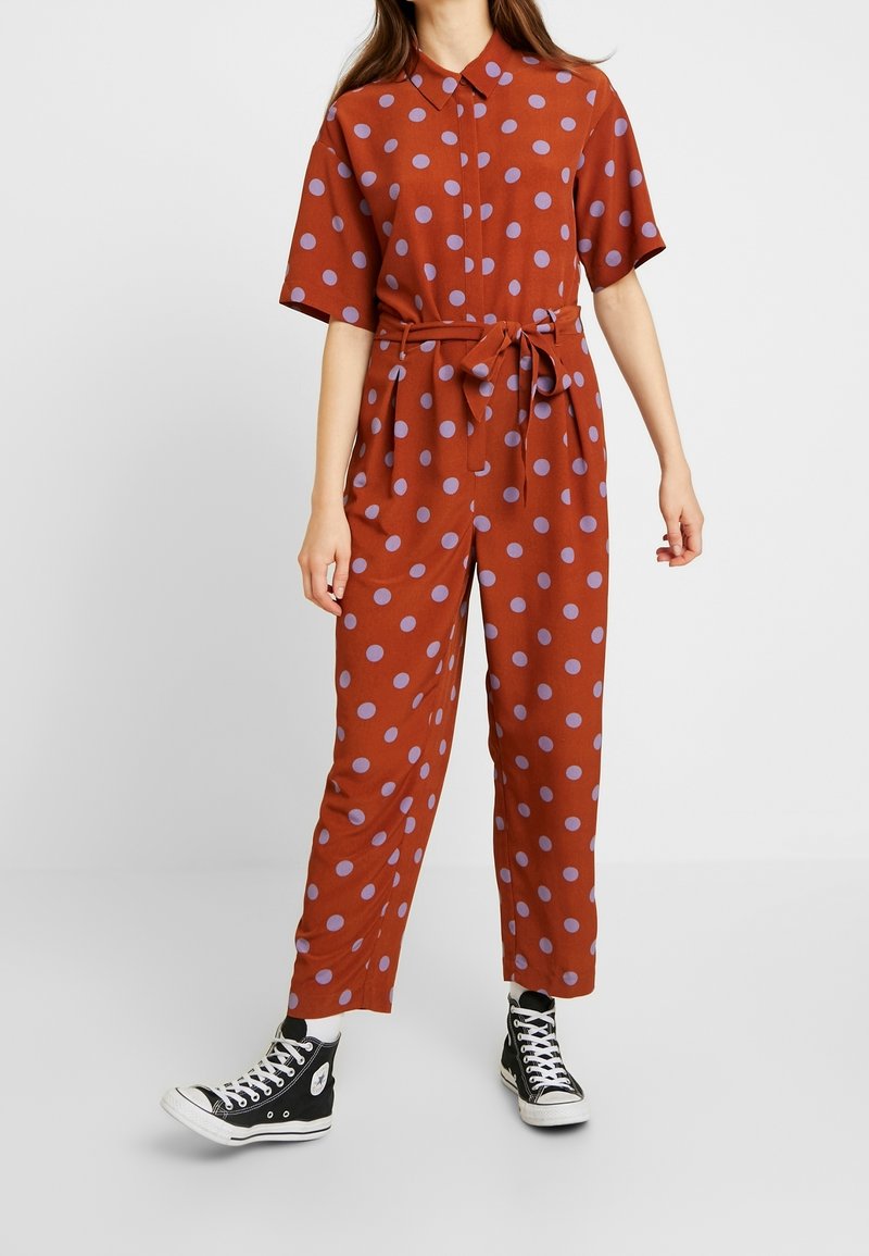 Personne portant une combinaison couleur rouille à pois violets, manches courtes, taille nouée, et baskets montantes noires sur fond blanc.