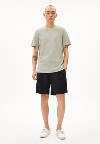 Hellgrünes, strukturiertes Rundhalsshirt kombiniert mit blauen Shorts, ergänzt durch weiße Schuhe. Das Modell hat Tattoos und einen rasierten Kopf.