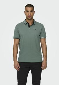 Grünes Polohemd aus Baumwolle, mit einem schwarzen Kragen, weißen und grünen Streifenakzenten sowie einem kleinen Logo auf der Brust.