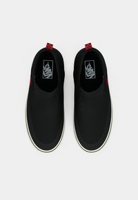 Black slip-on shoes with a smooth canvas upper, white rubber sole, red checkerboard accent at the heel, and Vans logo on the insole.