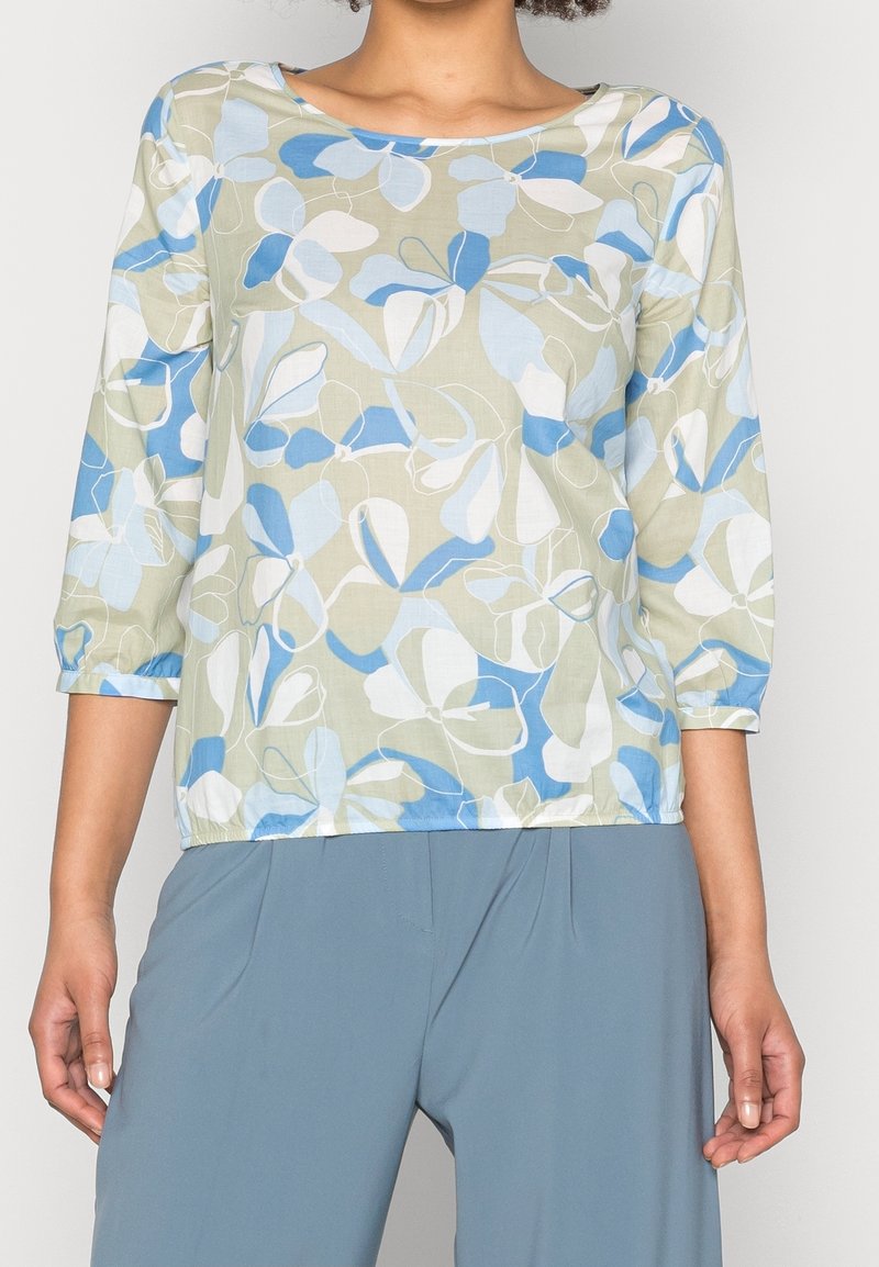 Femme portant un chemisier floral vert clair avec des feuilles bleues et blanches et des manches trois-quarts, associé à un pantalon bleu.