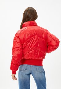 Rød pufferjakke med høj krave, kontrasterende rød ribkant og chevron-tekstildetalje. Båret over blå højtaljede jeans.