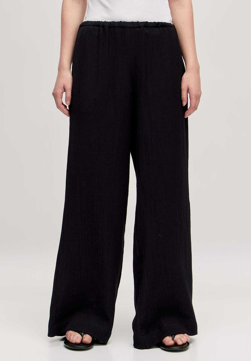Schwarze weit geschnittene Hosen aus strukturiertem Stoff, mit elastischem Bund und fließender Silhouette, kombiniert mit flachen Sandalen.