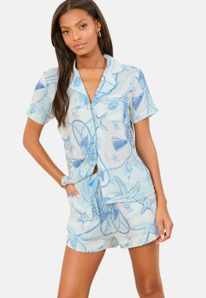 Žena nosi svetlomodrú košeľu s krátkym rukávom na gombíky a zodpovedajúce šortky s modrými vzormi mušlí a hviezdíc, stojí s jednou rukou v vrecku.