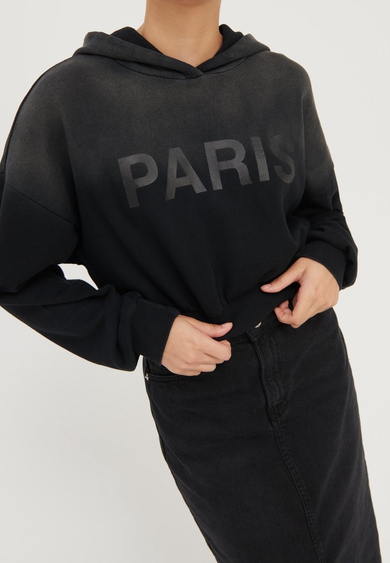 Sudadera negra con "PARIS" impreso en negro, cuenta con un bolsillo canguro y un corte relajado. Combinada con una falda de mezclilla negra.