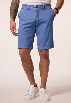 Mann trägt hellblaue Shorts mit aufgekrempeltem Saum, weiße Sneaker, weißes Hemd, mit tätowiertem Arm und schwarzer Armbanduhr, steht vor schlichtem Hintergrund.