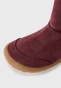 Gros plan sur une botte en daim rouge foncé avec des coutures détaillées et une semelle en caoutchouc marron clair sur un fond blanc.