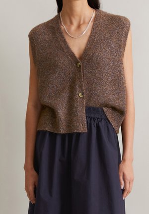 Ærmeløs brun strikket vest med knapper, båret over en sort nederdel med elastisk talje og en sølvkædehalskæde på en person.