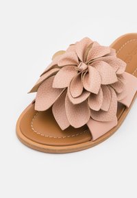 Sandale plate à bout ouvert avec une grande décoration fleur rose texturée sur la bride, sur une semelle marron avec des coutures blanches.