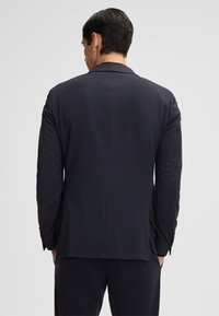 Giacca da abito blu navy con un sottile motivo testurizzato, spacco singolo sul retro e rever a punta. Presenta bottoni sulle maniche e tasche.