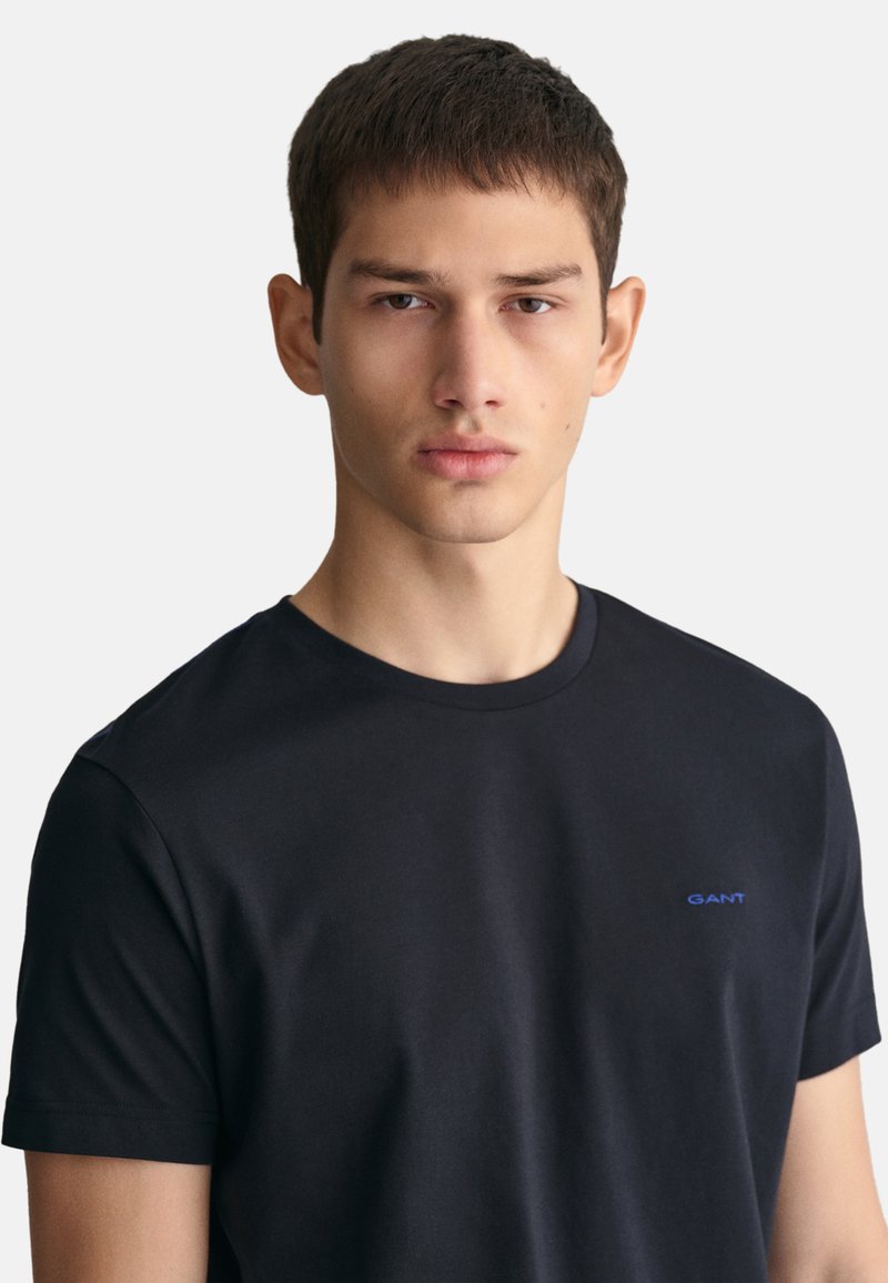Camiseta de algodón negra con cuello redondo y mangas cortas, con un pequeño logo azul de "GANT" en el pecho. Textura suave.