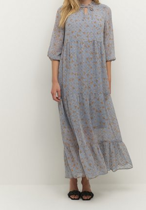 Femme portant une robe longue à manches longues bleu clair à motifs floraux avec un détail de nouage au col et des sandales noires tressées à bout ouvert, debout devant un fond blanc.