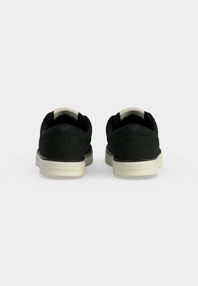 Paire de baskets basses noires avec semelles blanches, vues de dos, montrant le logo de la marque sur les languettes de talon et un texte en relief sur les semelles.