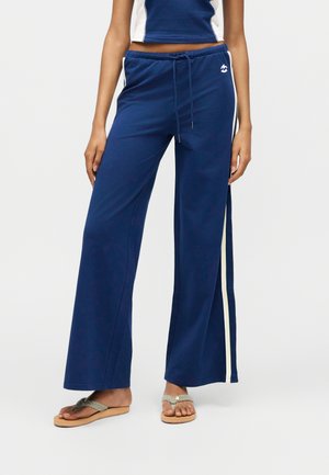 Γυναίκα που φοράει μπλε σκούρο παντελόνι με φαρδύ πόδι και λευκές πλαϊνές ρίγες, με κορδόνι στη μέση και σανδάλια με πλεγμένο λουράκι.