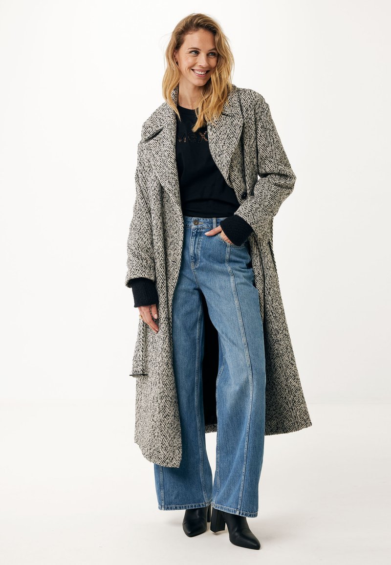 Houndstooth lang jasje in zwart en wit met brede revers, zwarte gebreide manchetten, gedragen over een zwart topje en hoge, wijde blauwe jeans.