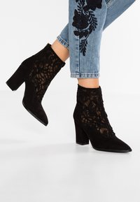 Botines negros de ante con tacón alto, que presentan un diseño de panel de encaje y una punta afilada. Superficie texturizada con un tacón grueso.
