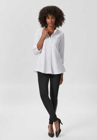 Witte button-up shirt met een gebogen zoom, lange mouwen opgerold tot de ellebogen, gecombineerd met zwarte leggings en puntige hakken.