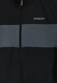 Sudadera con capucha negra con cremallera, con una franja horizontal gris en el pecho y un pequeño logo "FREDDY" en el lado izquierdo. Textura suave.