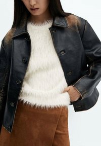 Czarna skórzana kurtka z lekko postarzanym wzorem, zapinana na guziki, noszona z białym, puszystym swetrem i brązową zamszową spódnicą.