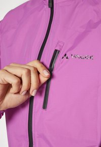 Giacca leggera di colore viola, realizzata in materiale liscio e resistente all'acqua. Presenta una cerniera nera e il logo Vaude sul petto.