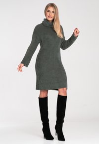 Strickpulloverkleid in Dunkelgrün, mit langen Ärmeln und einer lässigen Passform. Es hat eine gerippte Textur und einen Saum, der knapp über dem Knie endet.