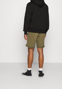 Persona che indossa un hoodie nero, pantaloni corti verde oliva arrotolati, calze bianche e sneaker nere alte, in piedi su uno sfondo bianco uniforme.