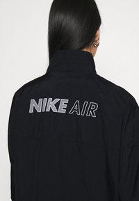 Svart jacka med hög krage, med texten "NIKE AIR" i vitt på baksidan. Slät struktur och lättviktsmaterial.
