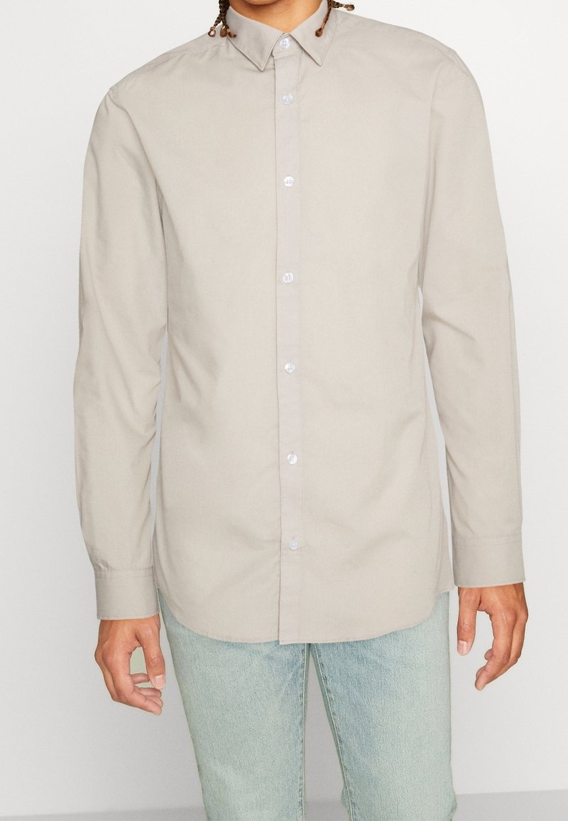 Personne portant une chemise beige clair à manches longues boutonnée avec col et un jean bleu clair, debout devant un fond uni.