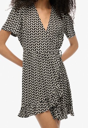Vrouw draagt een korte wikkeljurk met zwart-wit geometrisch patroon, korte mouwen en een gerimpelde zoom, poseert met handen achter haar rug.