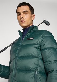 Man wearing dark green Calvin Klein puffer jacket holding a golf club over his shoulder against a plain background.