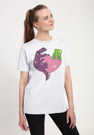 T-shirt blanc présentant un dinosaure violet de dessin animé et un ours vert, avec des détails en cœur rose. Manches courtes, col rond, coupe décontractée.