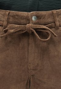 Pantalon en velours côtelé marron avec une ceinture en tissu nouée en nœud et une fermeture par bouton métallique à la taille.