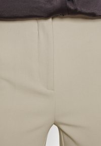 Pantalon beige avec une texture lisse, doté d'une poche latérale et d'une fermeture éclair invisible. Le matériau est léger avec des coutures minimales.