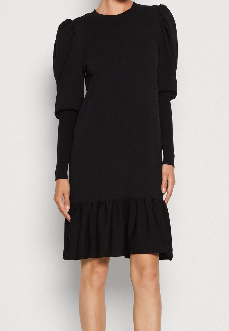 Robe noire jusqu'aux genoux avec manches longues bouffantes et ourlet volanté froncé, portée par une personne debout devant un fond uni.