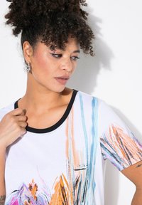 Camiseta blanca de manga corta con cuello negro, que presenta patrones abstractos coloridos en naranja, azul y rosa. Textura suave.