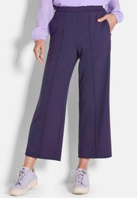 Gekürzte, weit geschnittene Hosen aus dunkelviolettem Stoff mit Seitentaschen, die über eine Mittelnaht verfügen und einen glatten Bund haben. Kombiniert mit hellvioletten Sneakers.