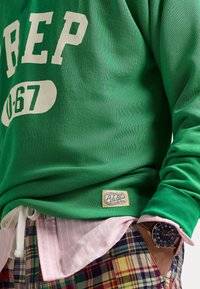 Zelená mikina s dlouhým rukávem s bílými nápisy a štítkem. Pod ní jsou vidět růžové pruhované tričko a kostkované šortky.