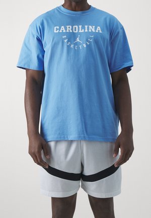 Mann trägt hellblaues Carolina Basketball-T-Shirt und weiße Netzshorts mit schwarzen Streifen, steht vor einem einfarbigen Hintergrund.