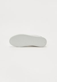 Hvit sneakersåle med teksturert gummi. Har jevnt fordelte horisontale spor. Rund tå og lett svikt i fotbeddet.