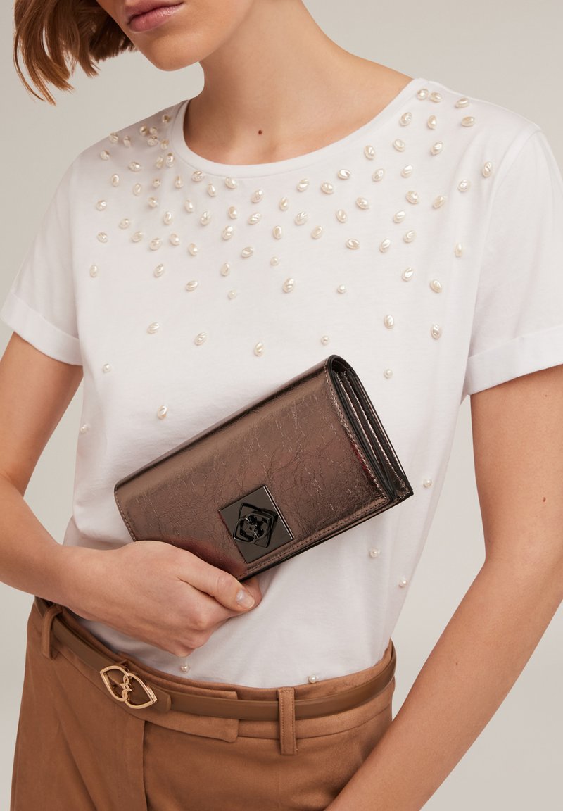 Metallisch braune Geldbörse mit einer strukturierten Oberfläche, ausgestattet mit einem schwarzen quadratischen Verschluss. Kombiniert mit einem weißen Hemd, das mit weißen, perlenartigen Verzierungen geschmückt ist.