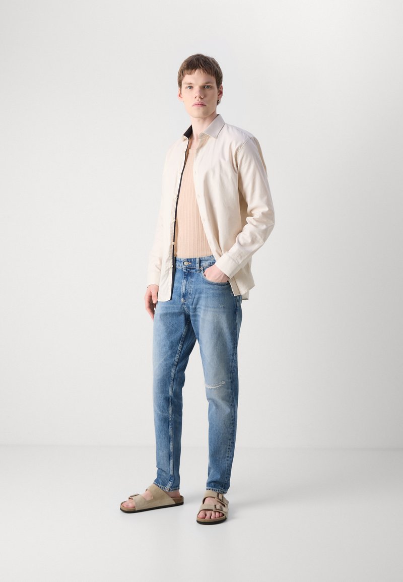 Hellbeige Hemd mit Knöpfen über einem gerippten beigen Tanktop, kombiniert mit ausgebleichten hellblauen Jeans und grauen Sandalen mit zwei Riemen.