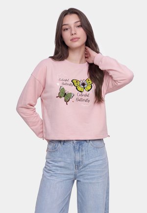 Ung kvinde med langt brunt hår iført en lyserød sweatshirt med farverige sommerfugletryk og lyseblå jeans, poserer mod en ensfarvet baggrund.