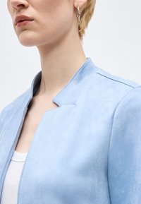 Άτομο που φοράει γαλάζιο σουέντ μπουφάν με γιακά με εγκοπές, λευκό τοπ από κάτω και μικρό ασημένιο κρίκο στο εμφανές αυτί.