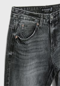 Jean en denim noir délavé avec poches avant, passants de ceinture, rivets métalliques, fermeture par bouton et étiquette de marque Stradivarius visible à l'intérieur de la ceinture.